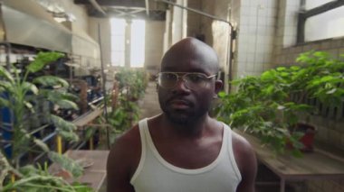 İş yerinde kameraya poz veren fanila ve gözlüklü Afrikalı genç fabrika işçisinin orta boy portresi.