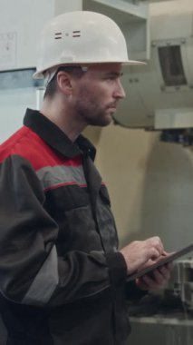 Arka planda çalışan CNC değirmen makinesini çalıştırmak için dijital tablet kullanan Kafkas erkek fabrika işçisinin dikey görüntüsü