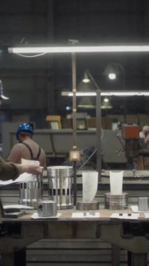 Beyaz tenli erkek metal fabrikası yöneticisinin dikey geniş portresi. Mavi şapkalı, çalışma masasına yaslanmış, lambaların altında makine detaylarıyla kamera önünde poz veriyor.