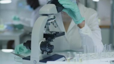 Beyaz laboratuvar önlüğü, yüz kalkanı, maske ve eldivenlerle laboratuvarda araştırma yaparken mikroskoptan bakan genç beyaz kadın bilim adamını yukarı kaldır.