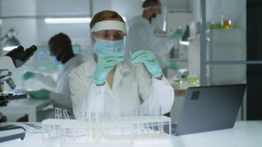Genç beyaz kadın mikrobiyolog koruyucu iş kıyafetleri içinde laboratuarda bilimsel araştırmalar yaparken test tüplerini incelerken ve dizüstü bilgisayara yazarken orta ölçekte çekilmiş.