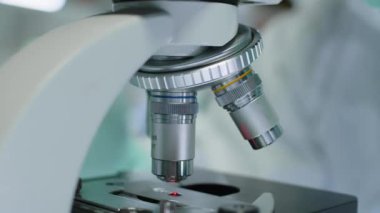 Laboratuvarda çalışırken mikroskopta dönen ve objektif lensleri değiştiren tanınamayan eldivenli bilim adamlarının yakın çekimi.
