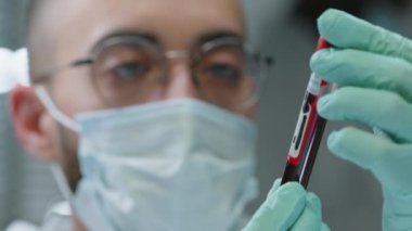 Gözlüklü, tek kullanımlık maske takan ve eldiven takan Orta Doğulu bir bilim adamının laboratuarda çalışırken elinde kırmızı sıvı bulunan test tüpüne baktığı yakın plan.
