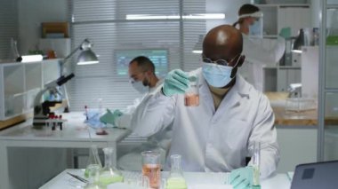Beyaz laboratuvar önlüğü ve yüz maskesi takmış Afro-Amerikan erkek kimyagerin orta yavaş portresi üzerinde cam eşyalar ile masada oturuyor ve kameraya bakıyor, laboratuvardaki meslektaşlarıyla çalışıyor.