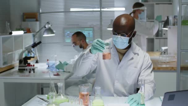 身穿白色实验室外套和面罩的非洲裔美国男性化学家的中慢幅画像 与实验室同事一起坐在桌旁 桌上放着玻璃器皿 看着相机 — 图库视频影像