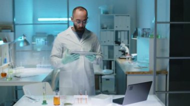 Modern laboratuarda çalışırken fotoğraf makinesine dijital tabletle poz veren laboratuvar önlüklü Ortadoğulu erkek bilim adamının orta boy portresi.