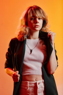 Şık ceketli genç kızın dikey görüntüsü neon arka planda duran kameraya bakıyor.