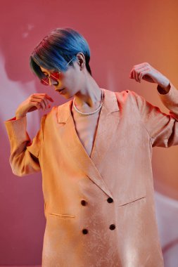 Şık ceketli, mavi saçlı, renkli arka planda kameraya poz veren genç adamın dikey görüntüsü.