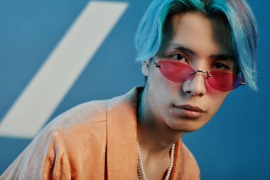 Pembe güneş gözlüklü ve mavi saçlı genç adamın portresi mavi arka planda kameraya bakıyor.