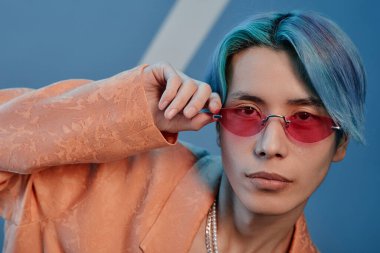 Mavi saçlı Asyalı bir gencin portresi. Mavi arka planda pembe güneş gözlüklü kameraya bakıyor.