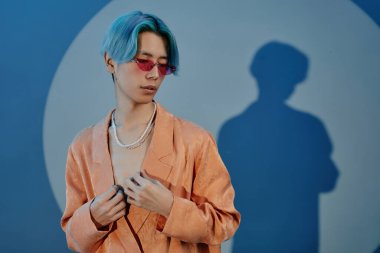 Ceketli Asyalı stil dansçısı portresi ve mavi arka planda kameraya poz veren pembe güneş gözlüğü.