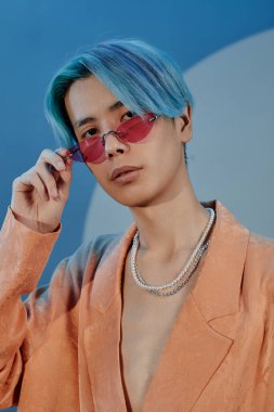 Mavi saçlı, şık gözlüklü, mavi arka planda kameraya bakan Asyalı genç adamın dikey görüntüsü.