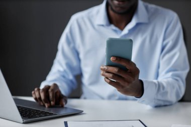 Ofisteki çalışma masasında otururken elinde akıllı telefon tutan tanınmayan siyahi işadamının yakın plan fotoğrafı.