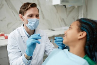 Diş sağlığı kliniğinde genç bir hastayla çalışırken maske ve eldiven takan arkadaş canlısı erkek dişçinin portresi.