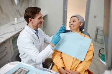 Gülümseyen erkek dişçinin, dişçi kliniğinde üst düzey bir kadınla çalışmasının yan görünümü..