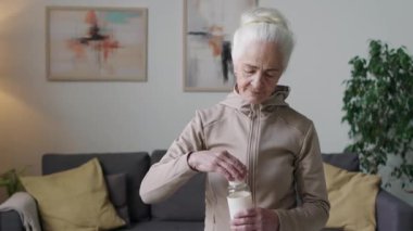 Modern bir dairede elinde spor şişesiyle kameraya poz veren fermuarlı kapüşonlu yaşlı kadının orta boy portresi.