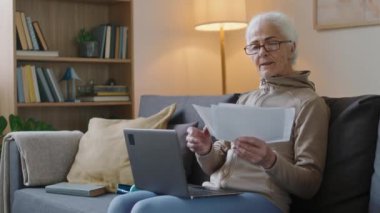 Kafkasyalı, gözlüklü yaşlı bir kadının oturma odasında oturmuş evrak işleriyle uğraşırken ve dizüstü bilgisayar kullanırken orta boy bir fotoğrafı.