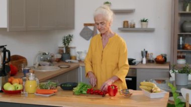 Orta boy beyaz, uzun hardallı elbise giyen, mutfakta yemek masasında duran ve evde öğle yemeği için salata yaparken taze sebze kesen yaşlı bir kadın.