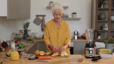 Orta boy, sağlıklı görünümlü, beyaz, yaşlı bir kadının evcil mutfakta vitamin kokteyli yaparken taze malzemeleri karıştırdığı bir fotoğraf.