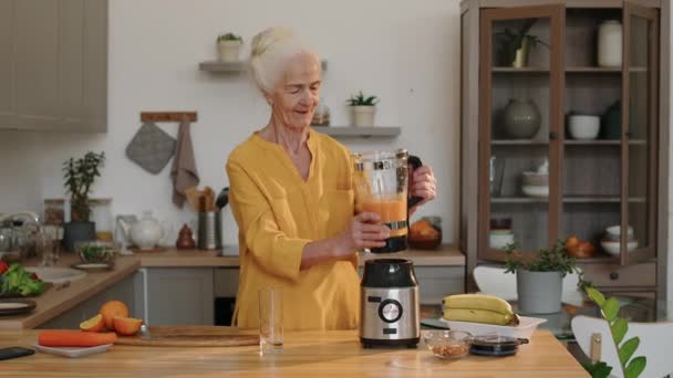 キッチンテーブルに立って ブレンダーからガラスに新鮮なビタミンスムージーを注ぐ 見栄えの良い白人老婦人の中型ショット — ストック動画