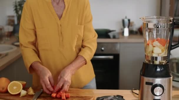現代のキッチンで朝食のためのビタミンスムージーを作っている間 オレンジとリンゴにロールカットニンジンを加える健康な白人高齢女性の中型ショットをトリミング — ストック動画