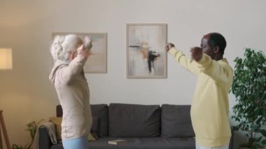 Etnik olarak farklı yaşlarda bir çiftin evde spor yaparken birbirlerinin önünde kol daireleri çizdiği orta ölçekli bir fotoğraf.