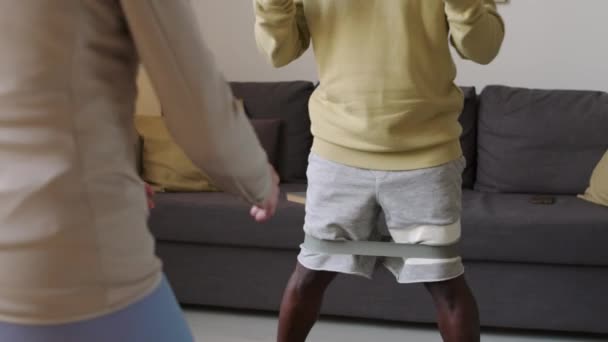 コーカサス女性コーチとの国内ワークアウトを持つ高齢の黒人男性のチルトショット 彼らの膝に抵抗バンドでサイドステップスカート運動 — ストック動画