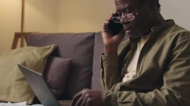 Yaşlı siyahi adamın kanepeye oturup cep telefonuyla konuşurken ve dizüstü bilgisayarında akşamları evden uzaktan çalışırken orta ölçekli bir fotoğrafını çekmek.
