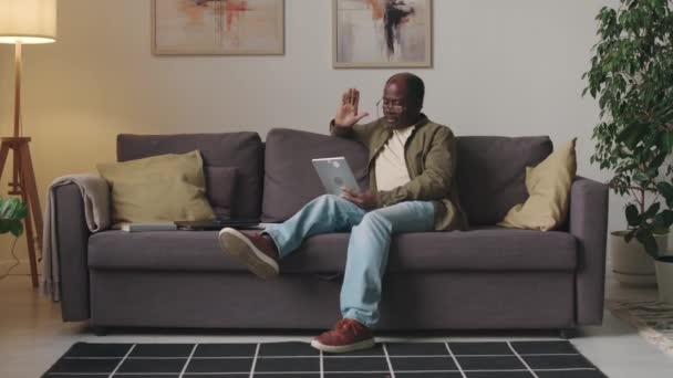 現代のリビングルームで快適なソファーに座って カジュアルな服やメガネのビデオチャットを身に着けているアフリカ系アメリカ人のシニア男性のフルショット — ストック動画