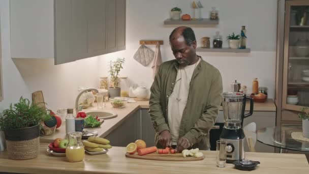 モダンなキッチンで料理テーブルに立って ブレンダーで健康的なスムージーを作っている間に木製のボードに新鮮なニンジンを切るアフリカ系アメリカ人のシニアショット — ストック動画