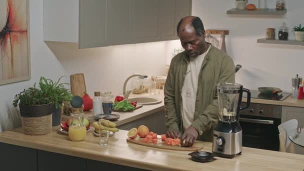 中等比例的退休黑人男子把新鲜胡萝卜和苹果放进搅拌机里 同时在现代化厨房里做早餐用的健康软糖 — 图库视频影像