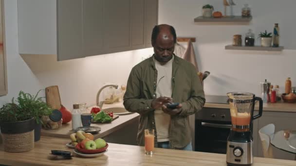 キッチンで調理テーブルで立っている間 スマートフォンと新鮮なニンジンスムージーのグラスを飲む大人の黒人の中型刺しショット — ストック動画