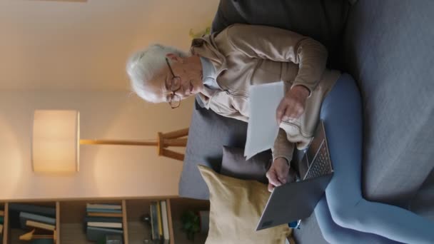 ラップトップチェック請求書付きリビングルームに座っているコーカサス高齢女性の垂直中型ショット — ストック動画