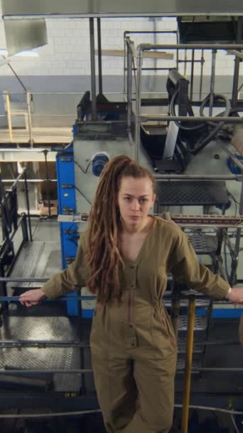 カーキのカバーオールのドレッドロックの若い白人女性の垂直な高角の広い肖像画は 背景に機械装置を備えた製造工場でカメラのためにリーニングし ポーズします — ストック動画