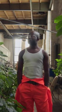 Dikey zoom portresi, beyaz atlet ve turuncu kaplama pantolon giyen yakışıklı zenci işçi fabrikada saksıda yeşil bitkilerin arasında dikilirken.