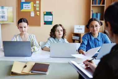 Bir grup okul çocuğu ders sırasında bilgisayar başında oturup öğretmenlerini dinliyorlar.
