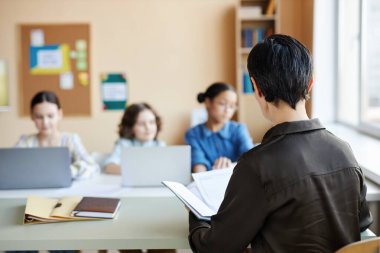 Öğretmeninin okulda otururken çocuklara not defterinde bir şeyler okuduğu arka görünüm