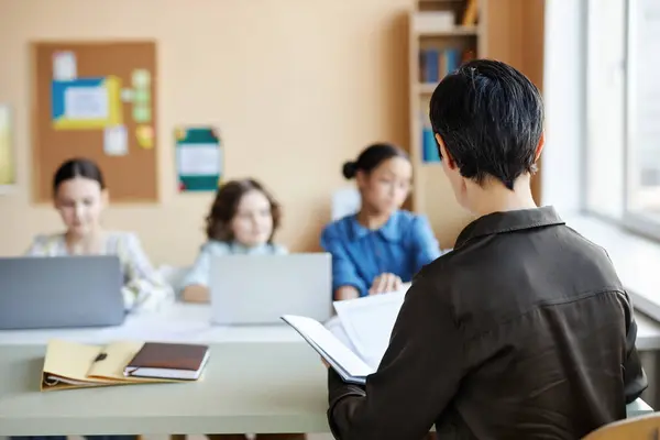 stock image Rear view of teacher reading something n her notebook to children while sitting at lesson at school