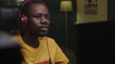 Akşamları kişisel bilgisayarda online video oyunu turnuvasına katılırken mikrofonlu oyun kulaklığı takan ve enerji içeceği içen genç Afrikalı Amerikalı adam.