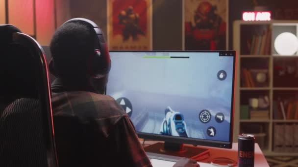 バックビューは 夜にシューティングゲームをプレイするネオンライトルームのデスクトップコンピュータの前に座っているマイクとチェッカーされたシャツとヘッドセットを身に着けている若い黒人男性ゲーマーのショットをウエスト — ストック動画