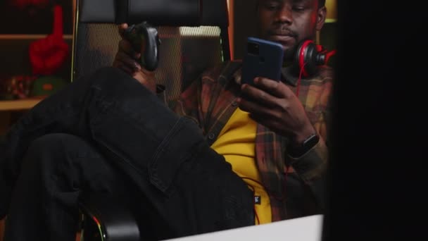 20대 초반의 아프리카계 미국인 남자가 스마트폰을 탐색하고 컨트롤러가있는 의자에 앉아있는 — 비디오