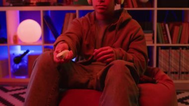 Orta ölçekli bir fotoğraf. Armut koltukta oturan, VR kulaklık takan ve sanal gerçeklik oyunları oynayan genç bir çocuk. Dairede neon kırmızı ışık yanıyor.