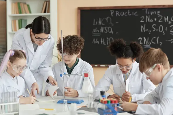 Diverse group of children wearing lab coats in science class at school with teacher helping