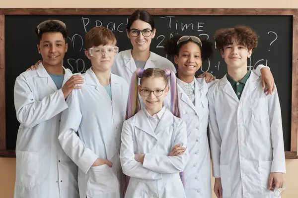 Diverse group of children wearing lab coats posing with teacher in science class at school and smiling at camera