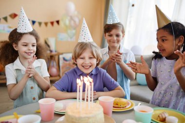Doğum günü partisinde dilek tutan mutlu küçük çocuğun portresi ve bir grup çocuğun tezahüratlarıyla pastaya mum üflemesi.