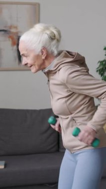 Kafkasyalı yaşlı bir kadının dambıllarla trisepsi ters çevirme egzersizi yaparken gündüz vakti evde spor yaparken dikey orta pozu.