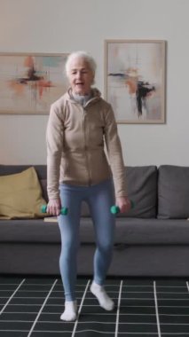 Aktif giyimli beyaz bir kadının evde dambıllarla çapraz atlama egzersizi yaparken dikey olarak tam bir bıçak darbesi.