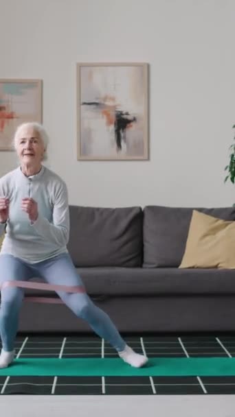 アクティブなコーカサス高齢女性の垂直刺しフルショット 彼女の膝に抵抗バンドは 昼間のホームワークアウト中にサイドステップスクワットを行います — ストック動画