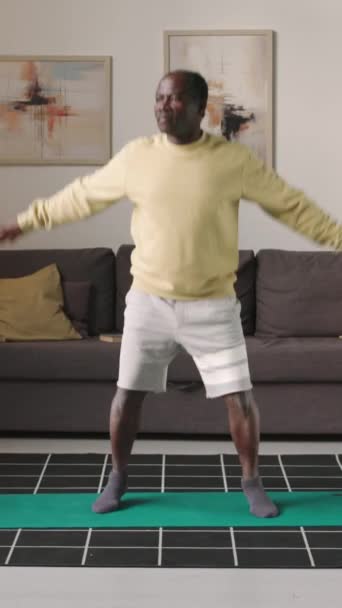 一名身穿运动服的黑人老年人白天在现代公寓的垫子上做跳跃运动时的垂直全刺镜头 — 图库视频影像