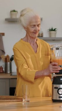 Mutfak masasının yanında duran ve blender 'dan taze vitamin içeceği döken beyaz bir kadının orta boy dikey fotoğrafı.